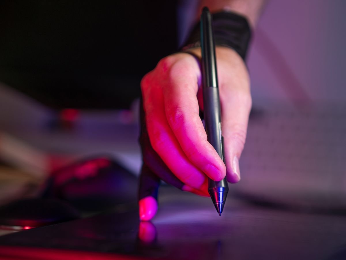 An artist's hand drawing with a stylus on a graphics tablet lit by pink studio light